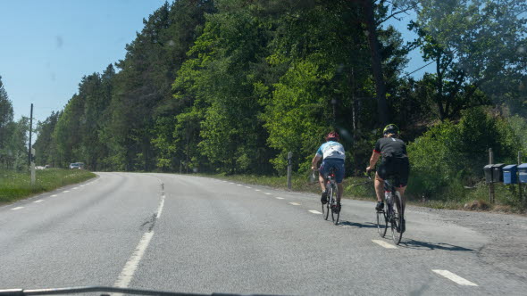 Två cyklister cyklar längs högerkanten av en landsväg. En bilist ska just köra om. En bit bort kommer en mötande bil.