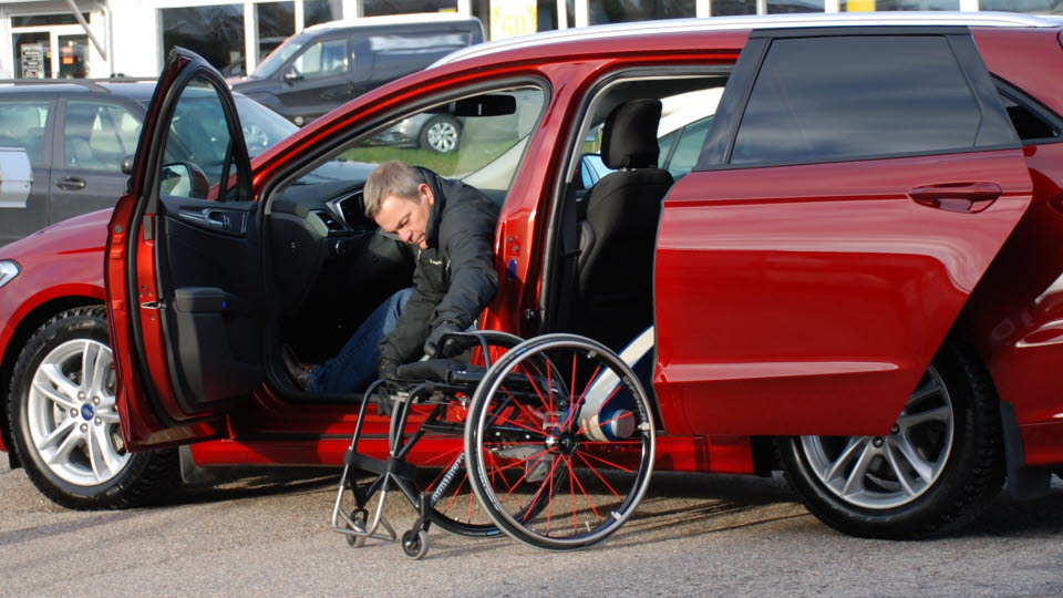 En man tar sig ur bilen, bredvid står en rullstol.