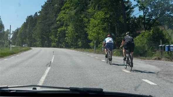 Två cyklister cyklar längs högerkanten av en landsväg. En bilist ska just köra om. En bit bort kommer en mötande bil.