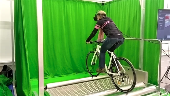 Cykelsimulator där en kvinna cyklar på rullar, i ett grönt hörn, med VR-glasögon på sig.