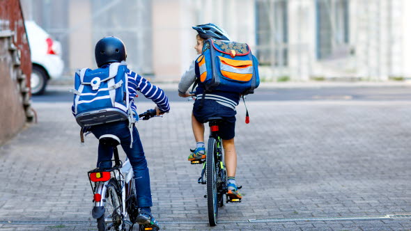 Två små skolpojkar cyklar tillsammans, med ryggsäckar och cykelhjälmar.