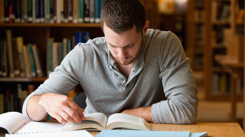 Ung man som sitter och läser på ett bibliotek.