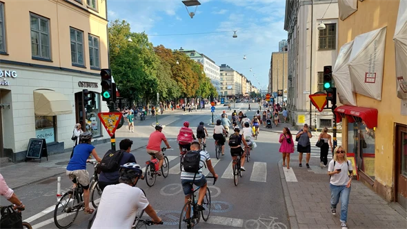 Många cyklister vid grönt ljus som cyklar längs Götgatan. På trottoaren går fotgängare. Inga bilar syns.