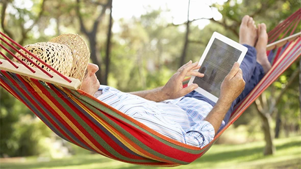 En man i stråhatt ligger i trädgården i en randig hängmatta och läser på en digital läsplatta.
