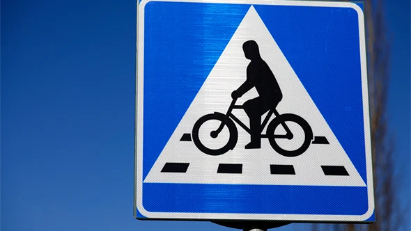 Trafikskylt Cykelöverfart (blå botten, vit liksidig triangel med svart figur som cyklar) mot blå himmel.
