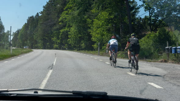 Två cyklister cyklar längs högerkanten av en landsväg. En bilist ska just köra om. En bit bort kommer en mötande bil.
