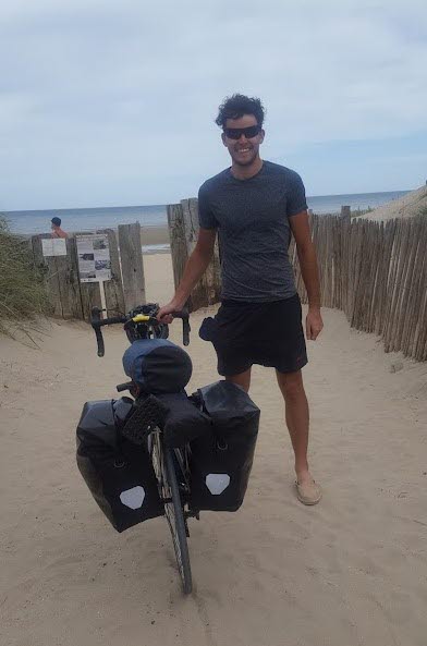 Foto på Kevin Gildea som står i shorts och t-shirt på en sandstrand med sin cykel.