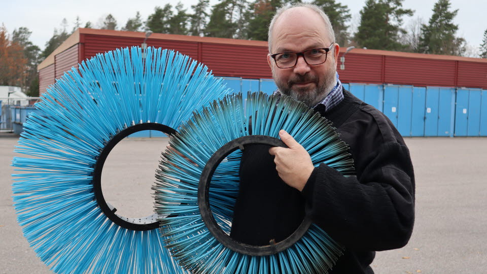 Göran håller upp två runda borstar utomhus med en industribyggnad i bakgrunden. 