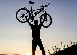 En person, som lyfter sin cykel upp i luften, syns i siluett mot soluppgången.
